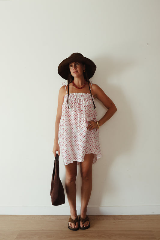 A white mini smock dress with a ruched bust and adjustable spagetti shoulder straps that tie. The fabric is 1a light weight 100% soft cotton in white with red polkadots.