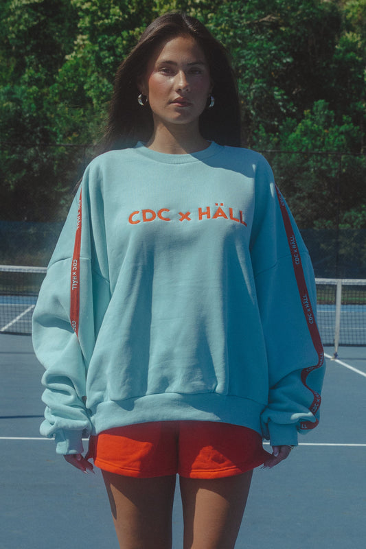 Person wearing a light blue sweatshirt with 'CDC x HALL' text on a tennis court