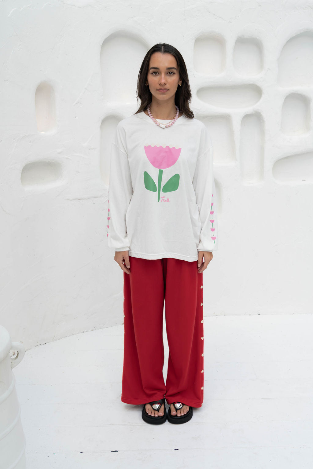 Women’s oversized long-sleeve t-shirt in white, featuring belle sleeves gathered into chunky cuffs. Designed in ultra-soft 100% organic cotton, it showcases a bold flower graphic in pink and green on the front and repeated down each sleeve, with a small 'häll' logo detail.