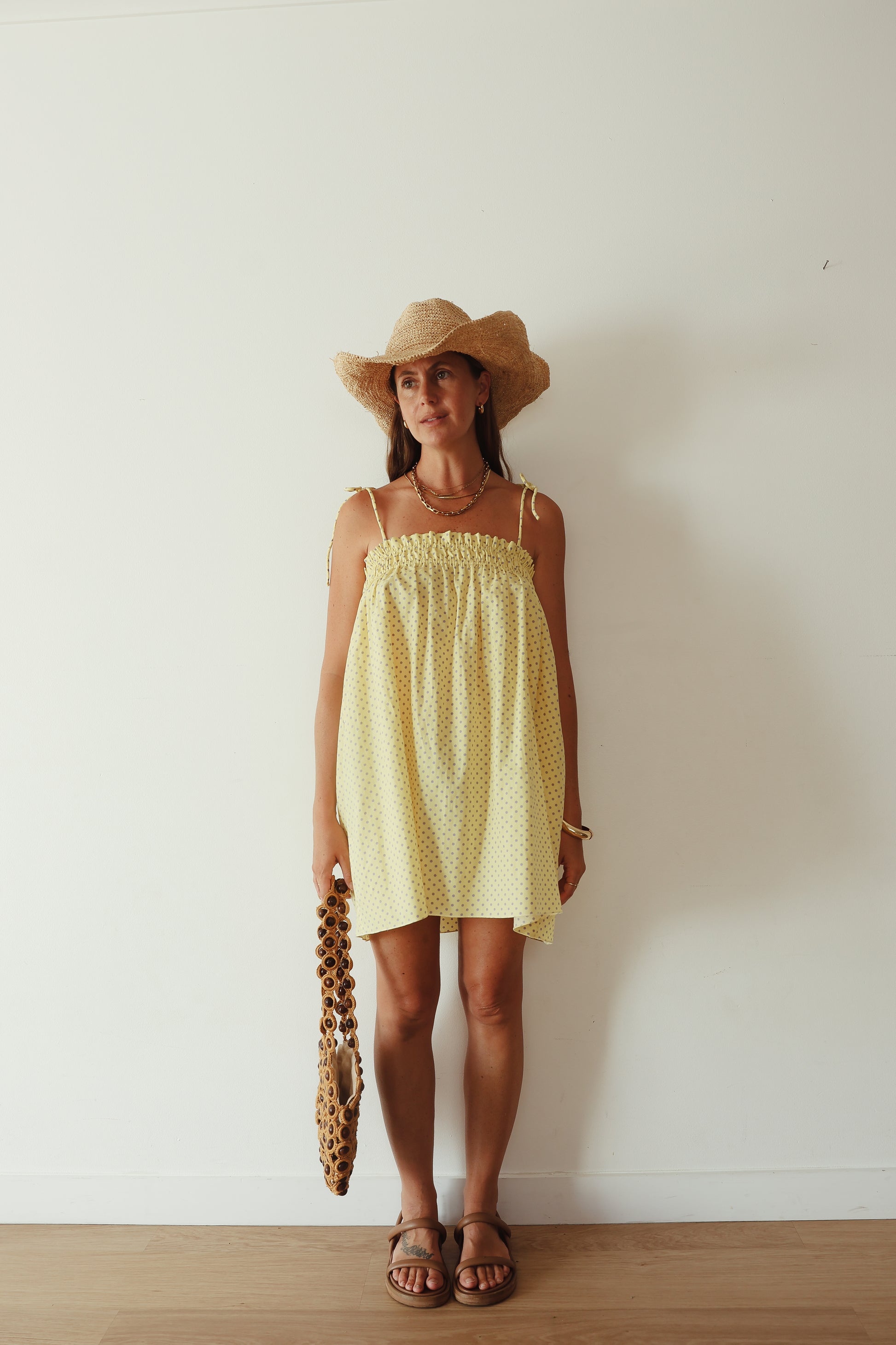 A yellow mini smock dress with a ruched bust and adjustable spagetti shoulder straps that tie. The fabric is a light weight 100% soft cotton in butter yelllow with purple lilac polkadots.