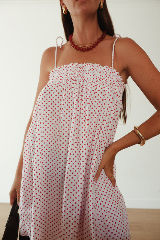 A white mini smock dress with a ruched bust and adjustable spagetti shoulder straps that tie. The fabric is 1a light weight 100% soft cotton in white with red polkadots.
