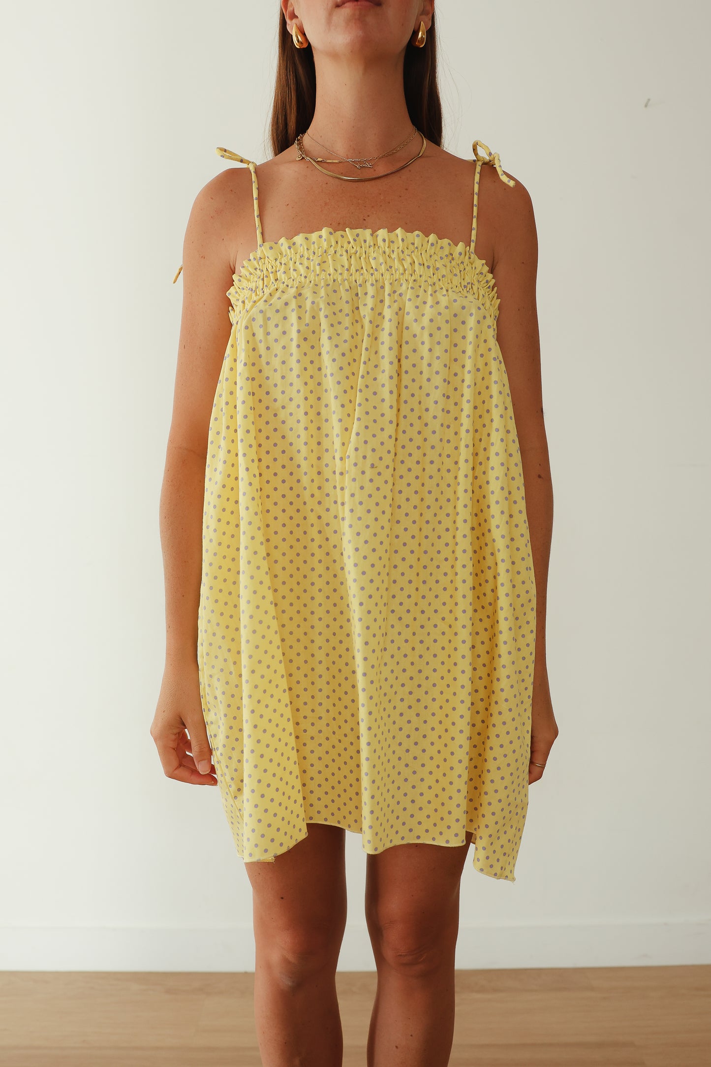 A yellow mini smock dress with a ruched bust and adjustable spagetti shoulder straps that tie. The fabric is a light weight 100% soft cotton in butter yelllow with purple lilac polkadots.