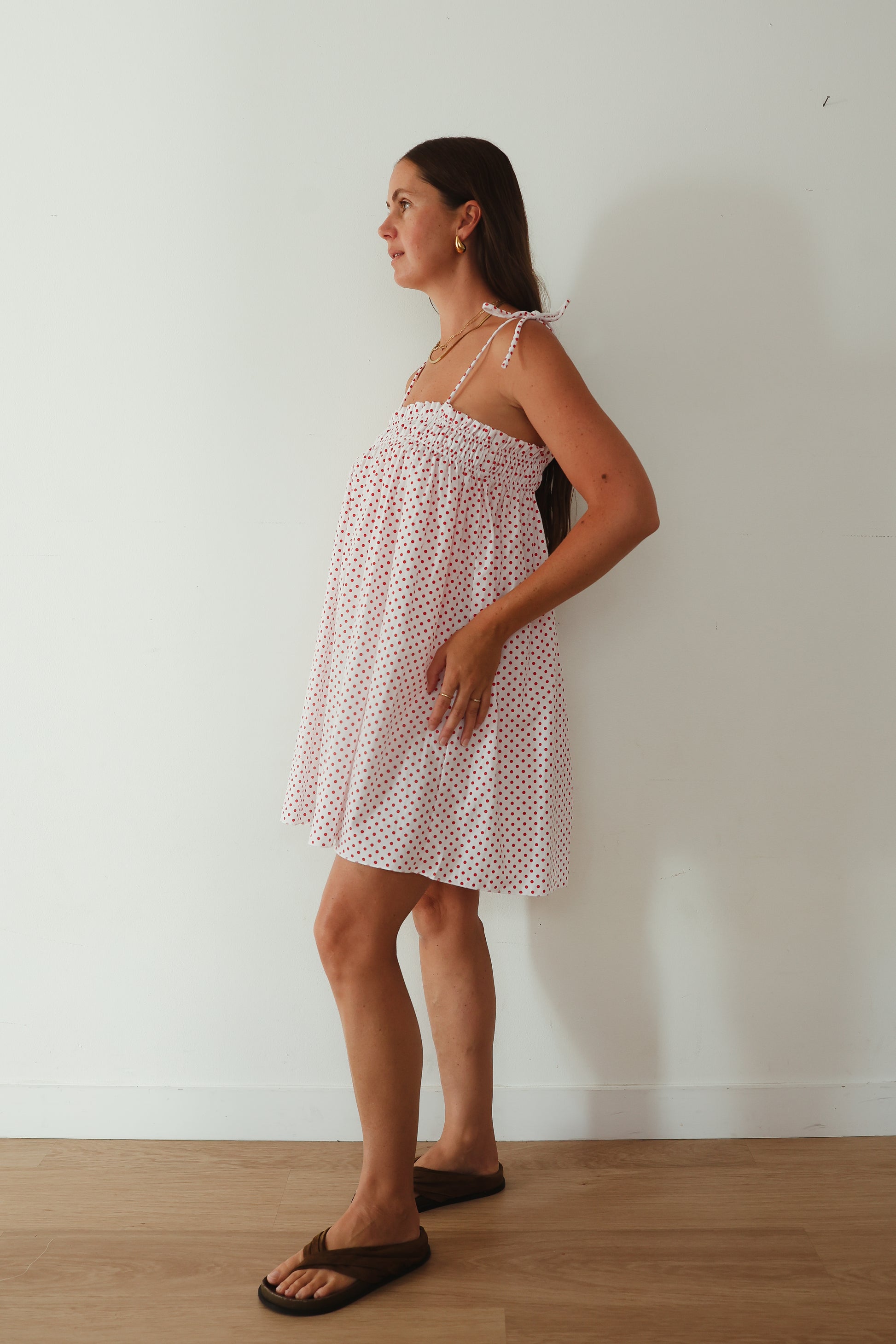 A white mini smock dress with a ruched bust and adjustable spagetti shoulder straps that tie. The fabric is 1a light weight 100% soft cotton in white with red polkadots.
