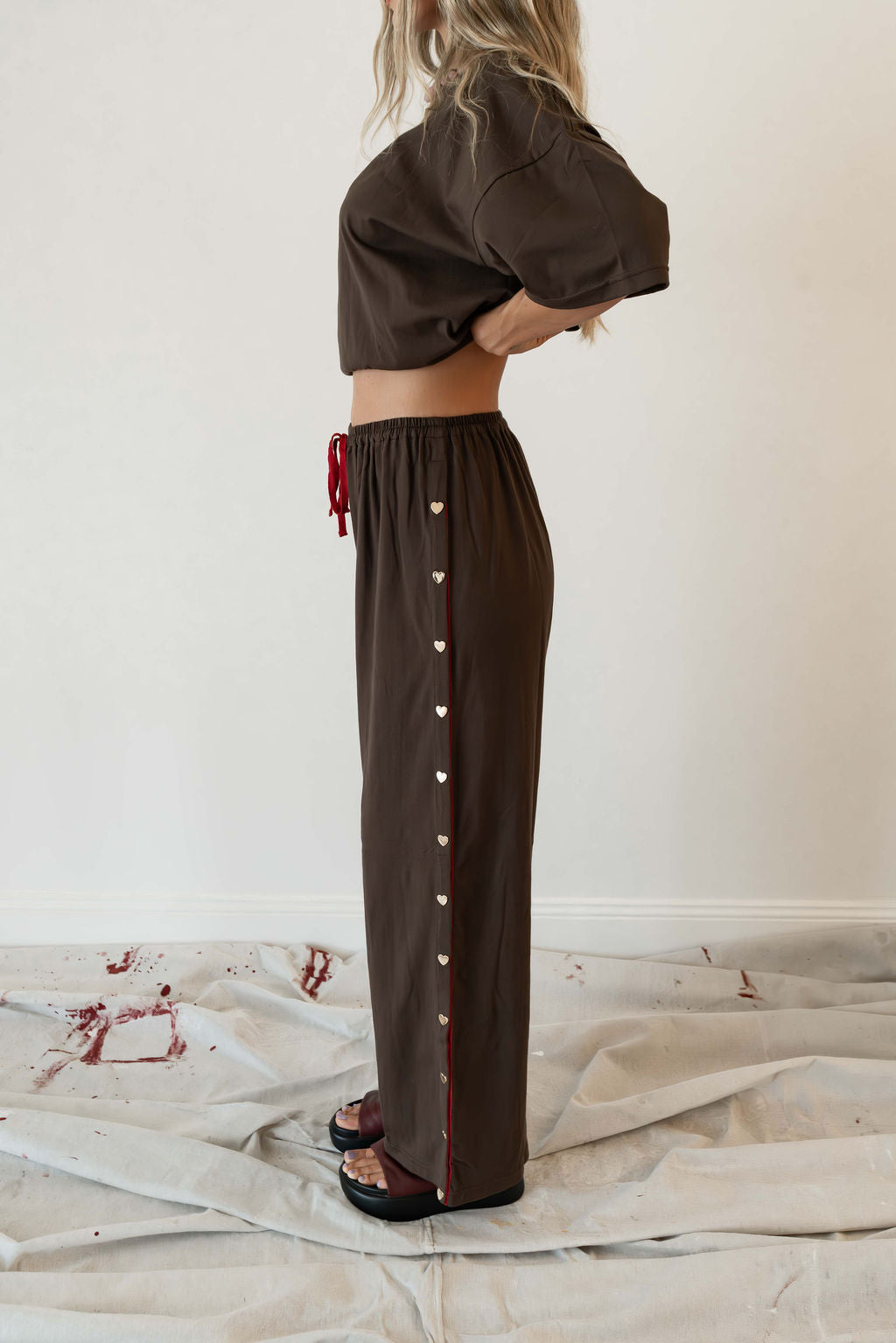 Model wears a two piece set in a rich chocolate brown organic cotton relaxed wide leg pant with side gold heart snap buttons down the leg with an oversized tshirt also in the rich chocolate brown organic cotton