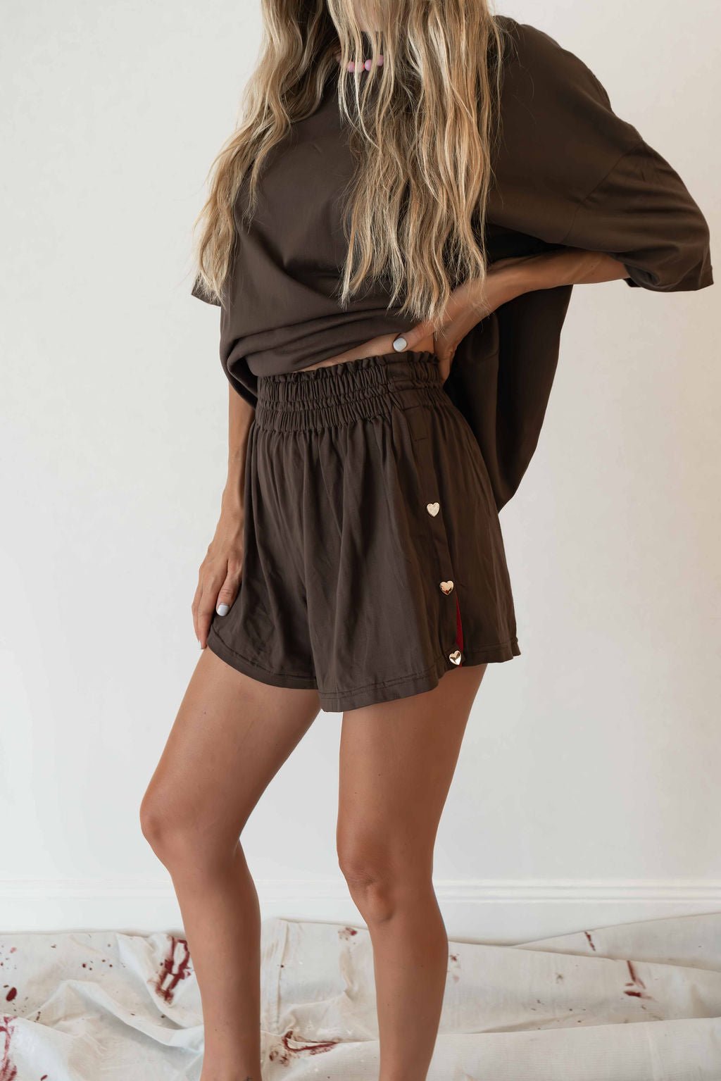 Model wears a two piece set in a rich chocolate brown organic cotton relaxed short with side gold heart snap buttons down the leg and a contrast red stripe down side with an oversized tshirt also in the rich chocolate brown organic cotton