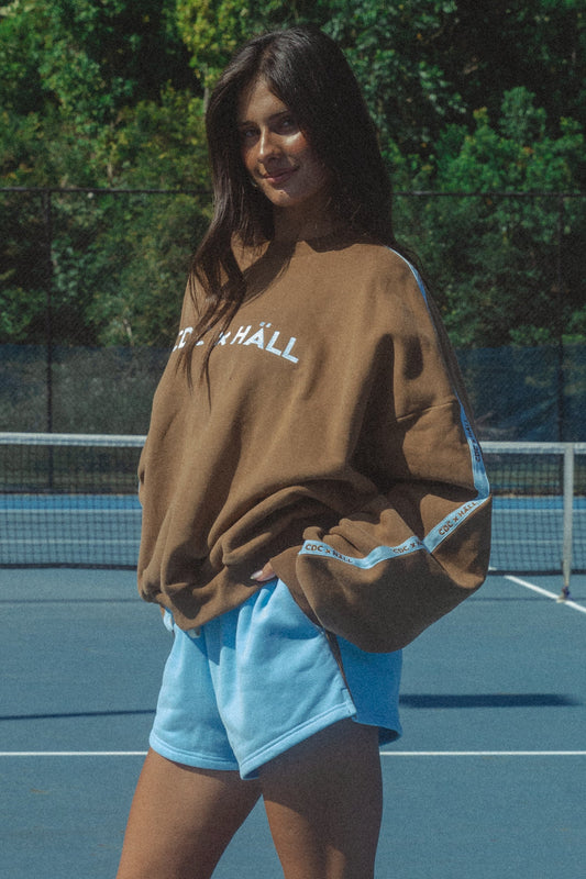 Woman wearing a brown sweatshirt with 'cracker day club x HALL' text on a tennis court. Sweater is cloud soft.
