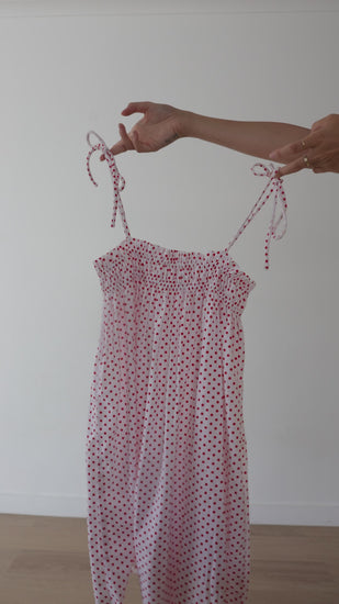 A white mini smock dress with a ruched bust and adjustable spagetti shoulder straps that tie. The fabric is 1a light weight 100% soft cotton in white with red polkadots.